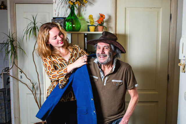 Een jonge vrouw staat naast een zittende man en helpt hem een jas aantrekken