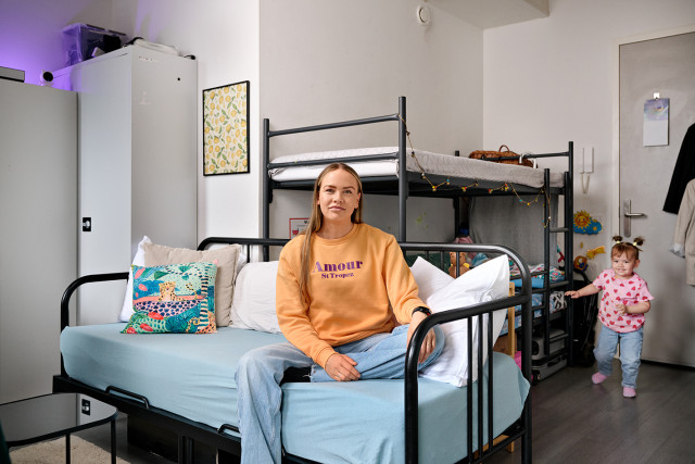 Vrouw zit op een bank, kindje komt naar haar toe lopen vanaf een stapelbed