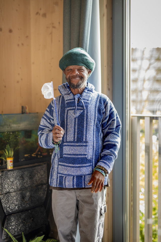 Man met blauwe trui en groene muts staat glimlachend in zijn woning